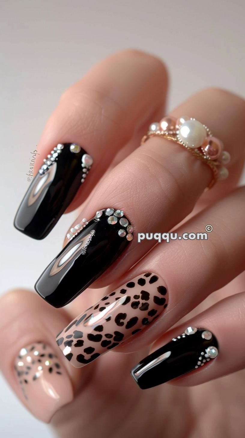 Stylish long acrylic nails with black polish adorned with rhinestones and nude polish with black leopard print pattern.