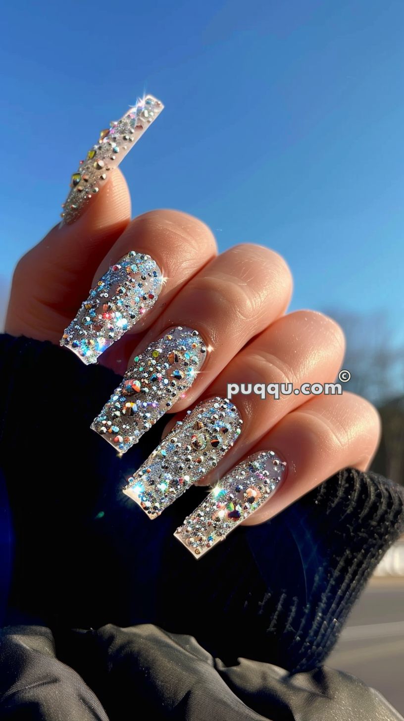Hand with long nails adorned with glitter and multicolored rhinestones, set against a clear blue sky.