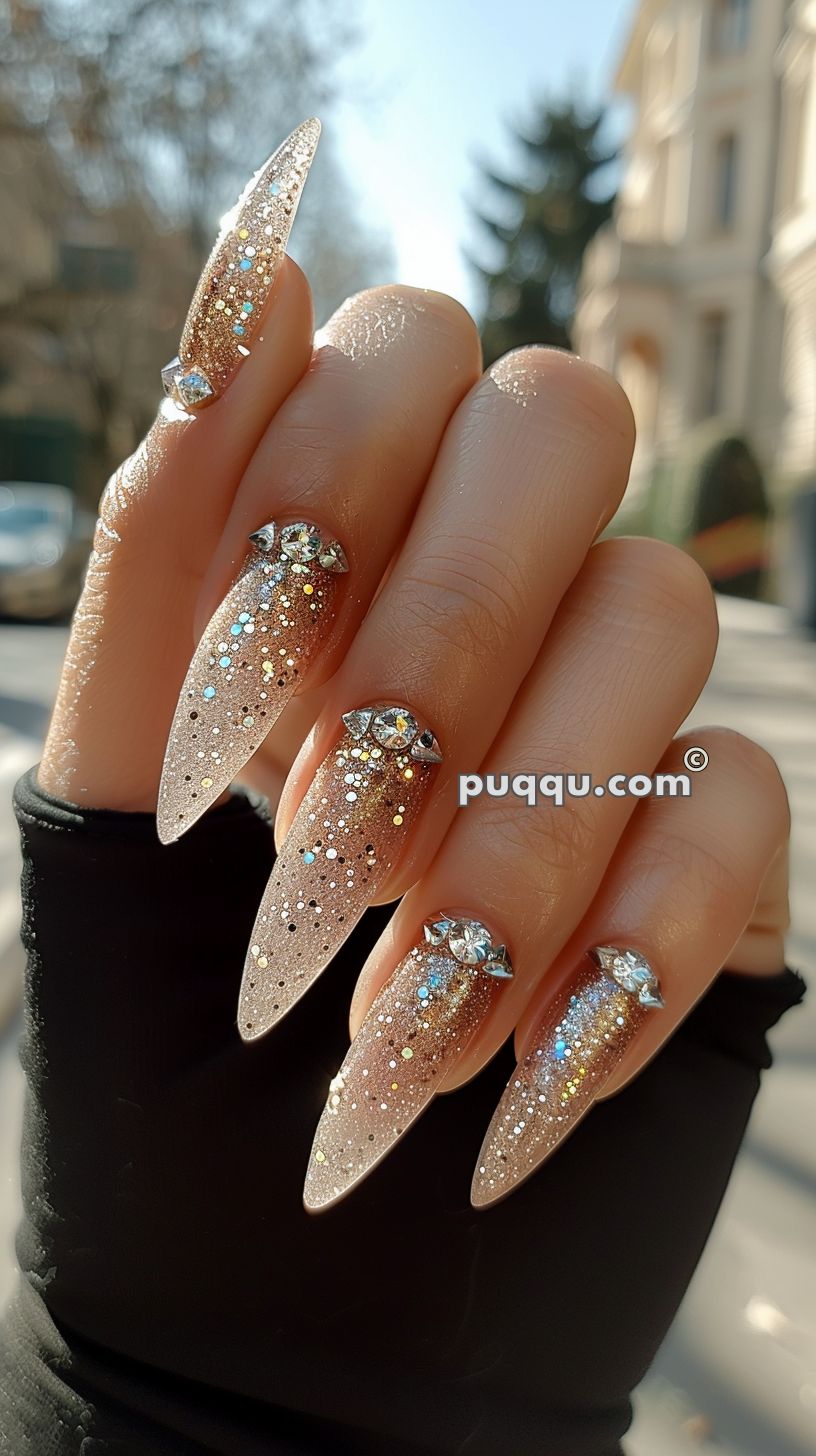 Hand with long, pointed nails decorated with shimmering gold glitter and adorned with rhinestones, against an outdoor background.
