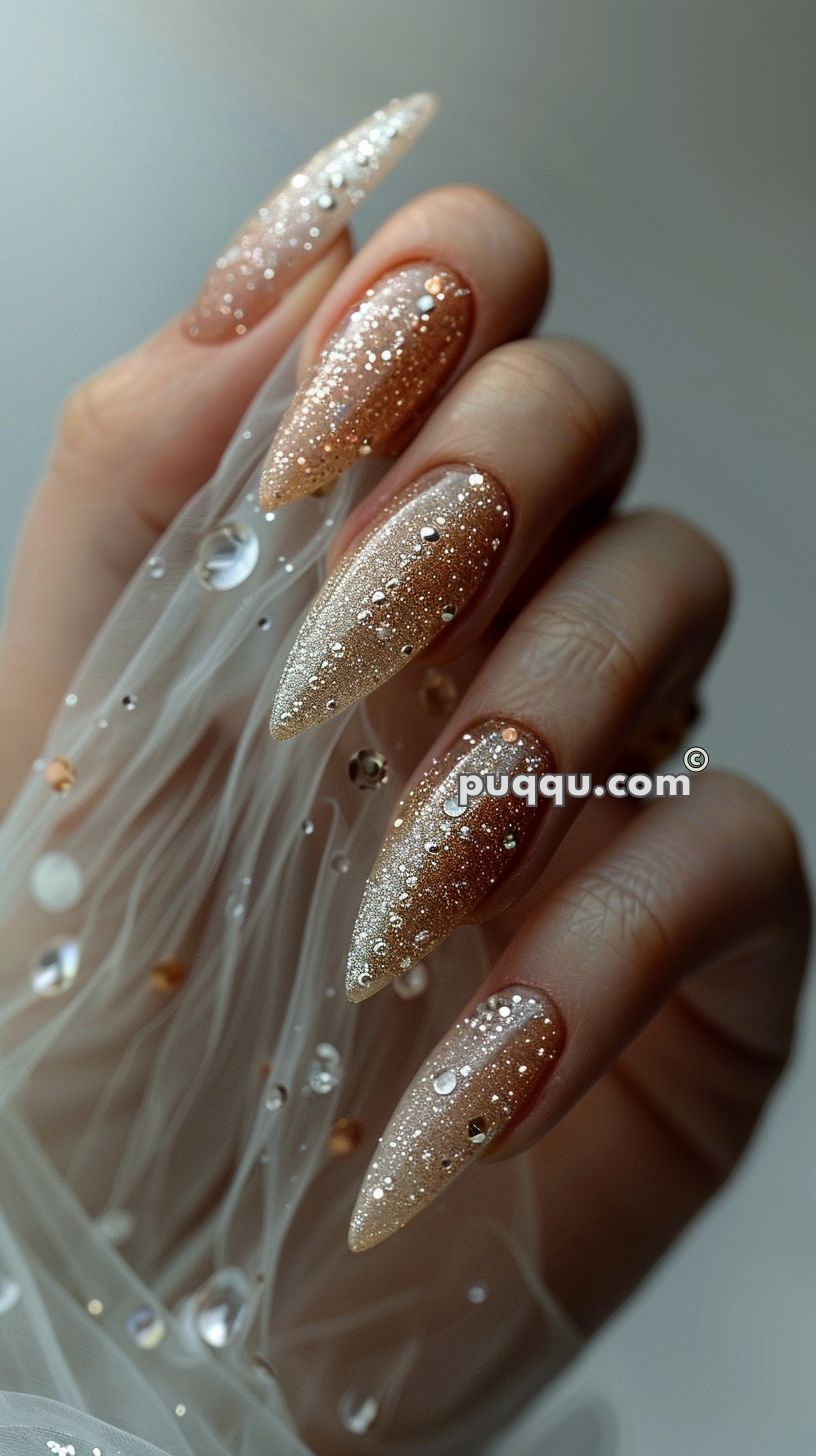 Long, almond-shaped nails with a gradient glitter design, embellished with small rhinestones, holding a sheer fabric with scattered embellishments.