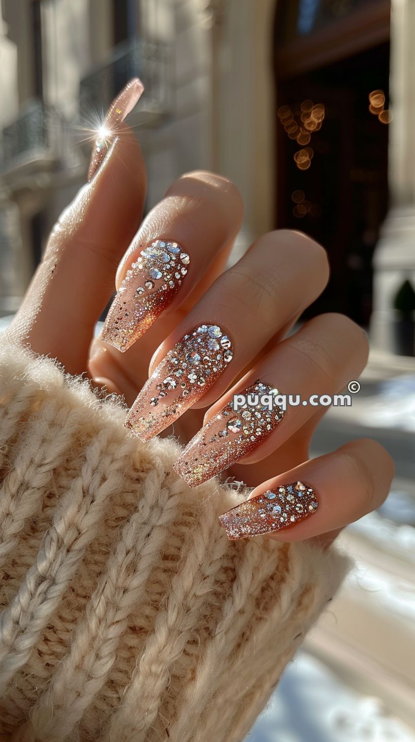 Long, sparkly gold nail art with rhinestones on fingers wrapped in a beige knitted sweater.
