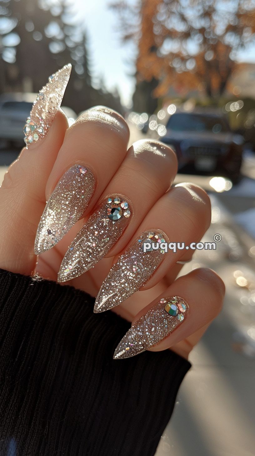 Close-up of hand with long, pointed silver glitter nails embellished with rhinestones, held against an outdoor background with trees and parked cars.