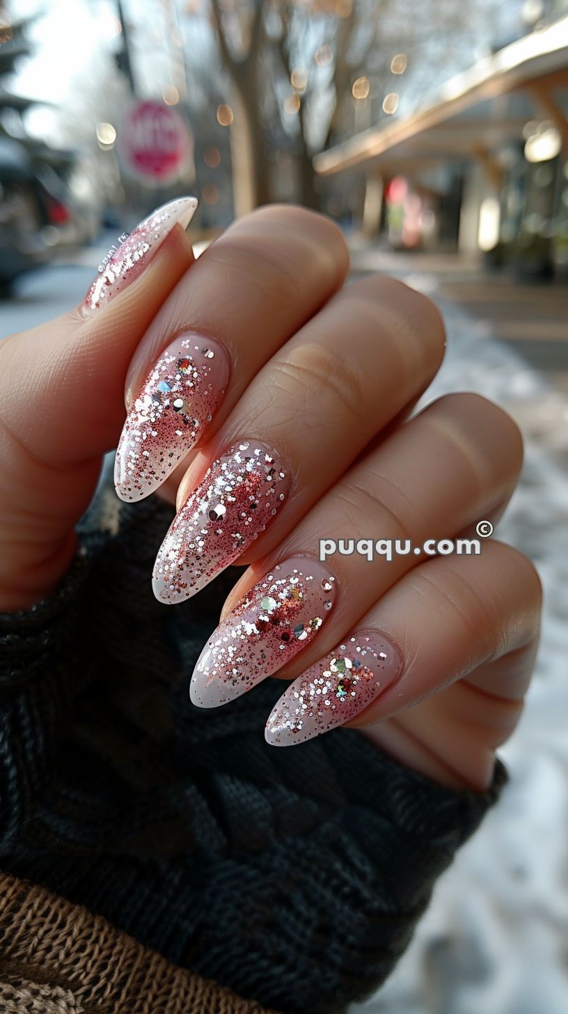 A hand featuring long, almond-shaped nails with a gradient pink and white gel polish, topped with chunky and fine silver glitter. The background shows an outdoor winter scene with bare trees and blurred buildings.