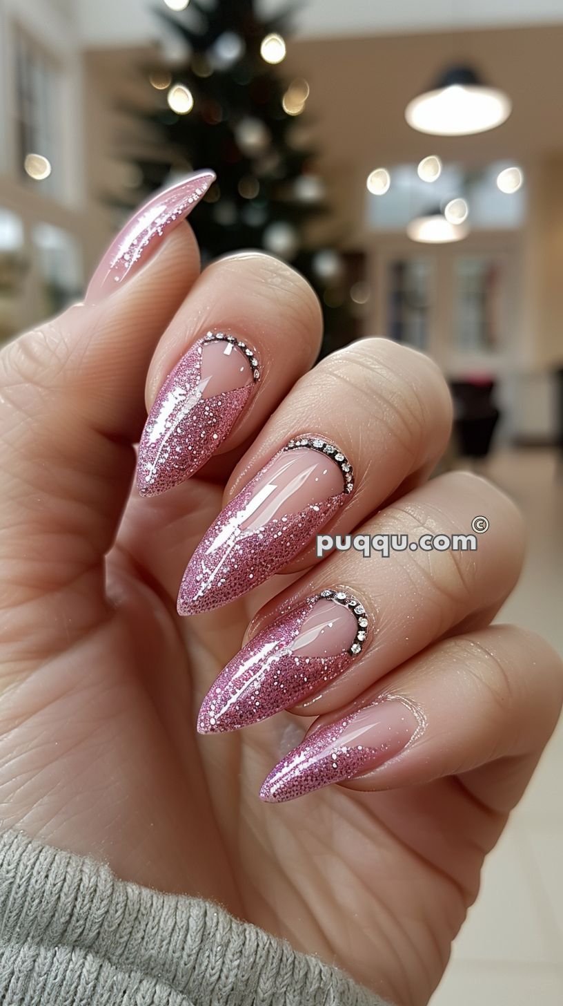 Hand with long, pointed nails featuring pink glittery polish and rhinestone accents near the cuticles, against a blurred background with Christmas tree lights.