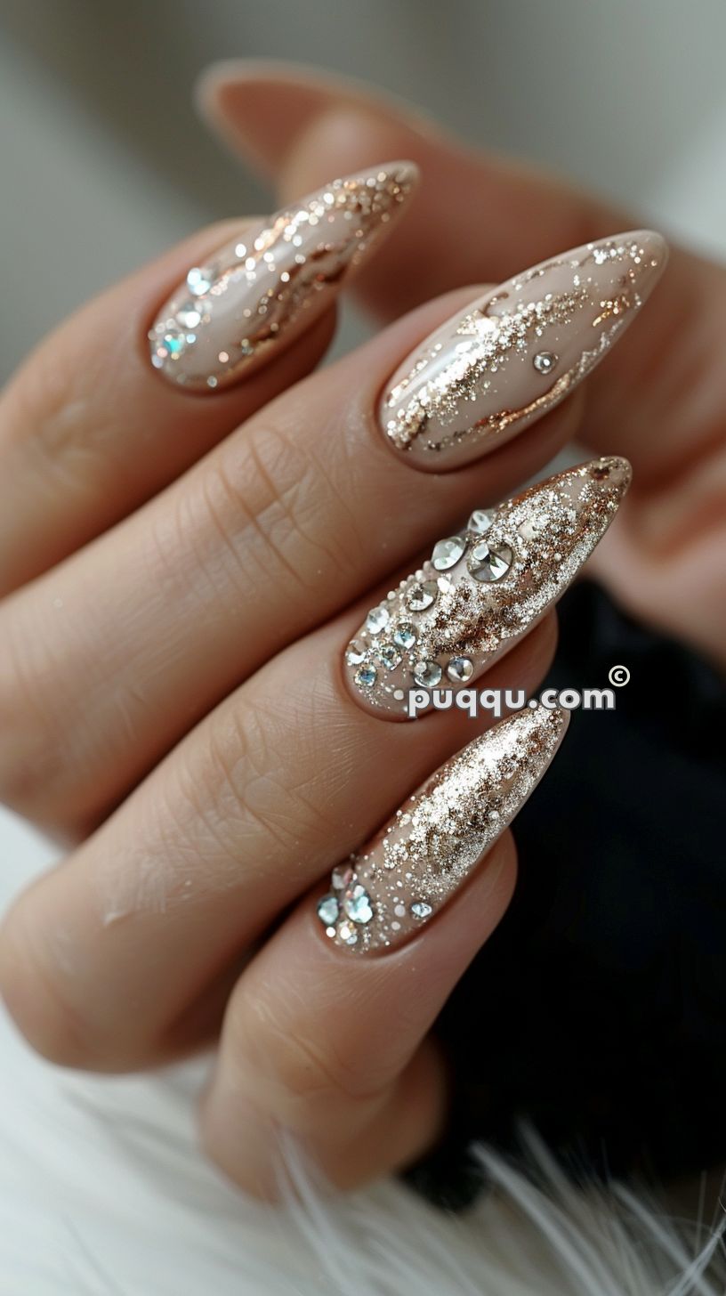 Close-up of a hand with long, pointed nails decorated with a beige base, gold glitter, and rhinestone embellishments.