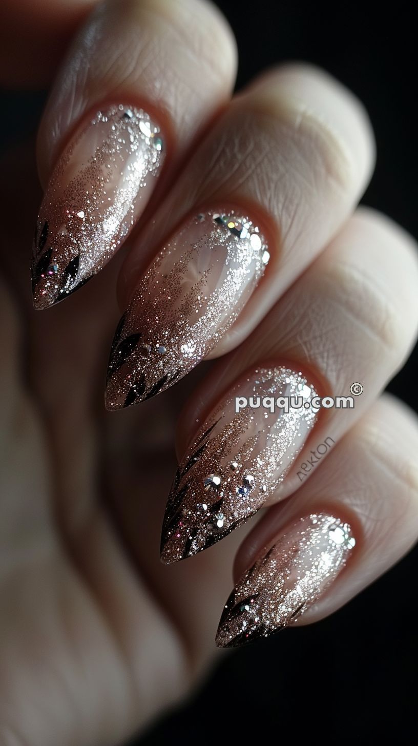 Close-up of a hand with almond-shaped nails featuring a glittery gradient design, starting with a nude base that blends into silver glitter with black accent lines and small rhinestones.