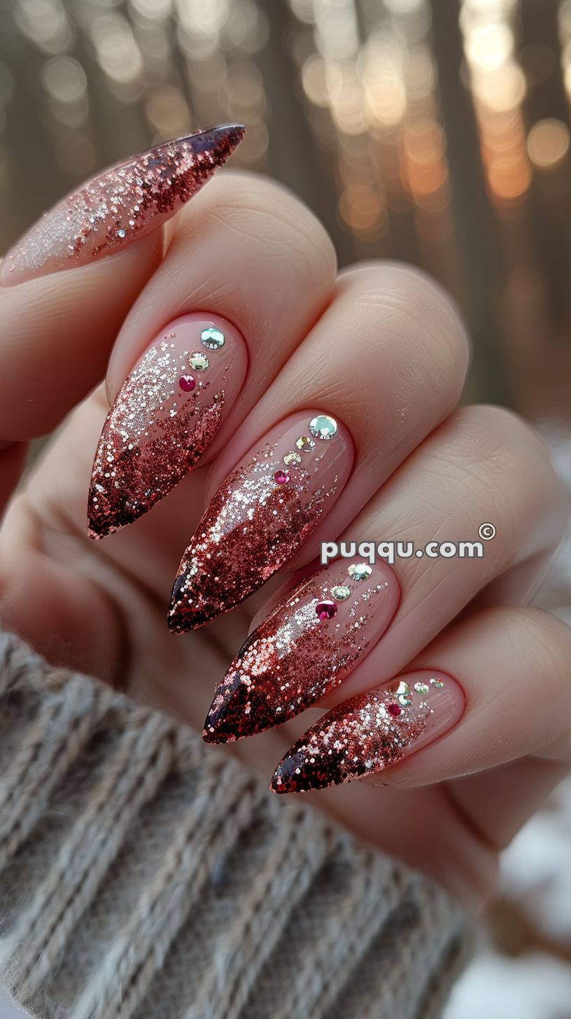 Close-up of pointed nails with pink and black glitter ombre design, adorned with rhinestones and red heart accents, with a blurred background.