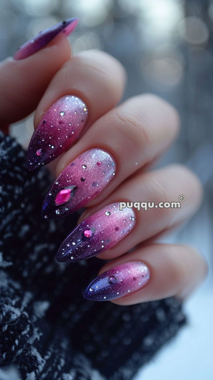 Close-up of a hand with long stiletto nails featuring a purple to pink gradient with glitter and rhinestone decorations.