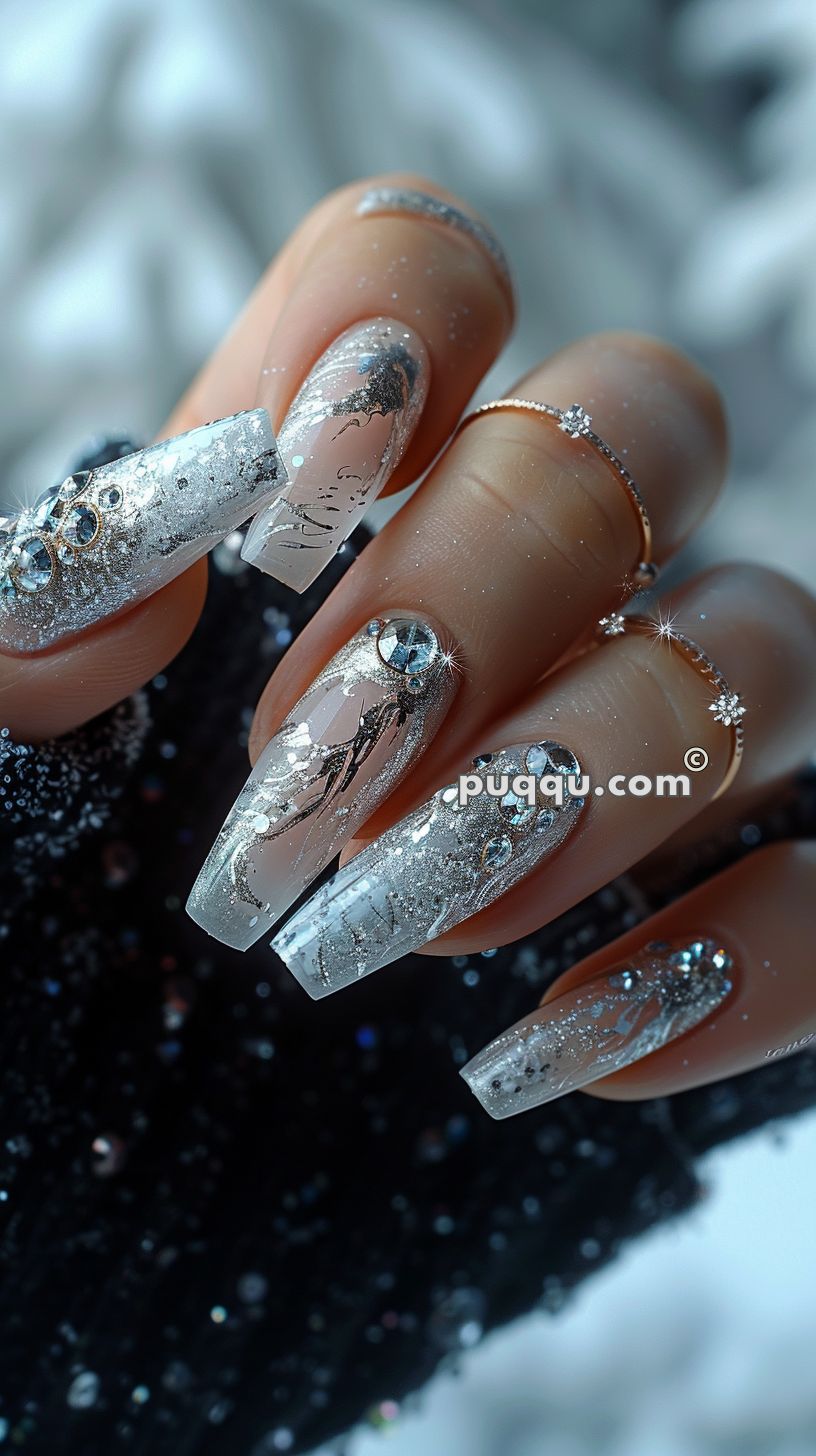 Close-up of a hand with long, square-shaped nails featuring intricate silver and clear designs, adorned with rhinestones and glitter. The person is wearing delicate rings and a black glittery glove on one hand.