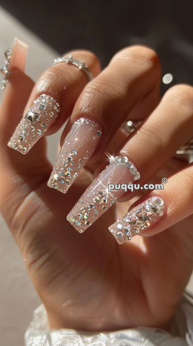 Close-up of a hand with long, square-shaped nails decorated with pink glitter and various rhinestones.