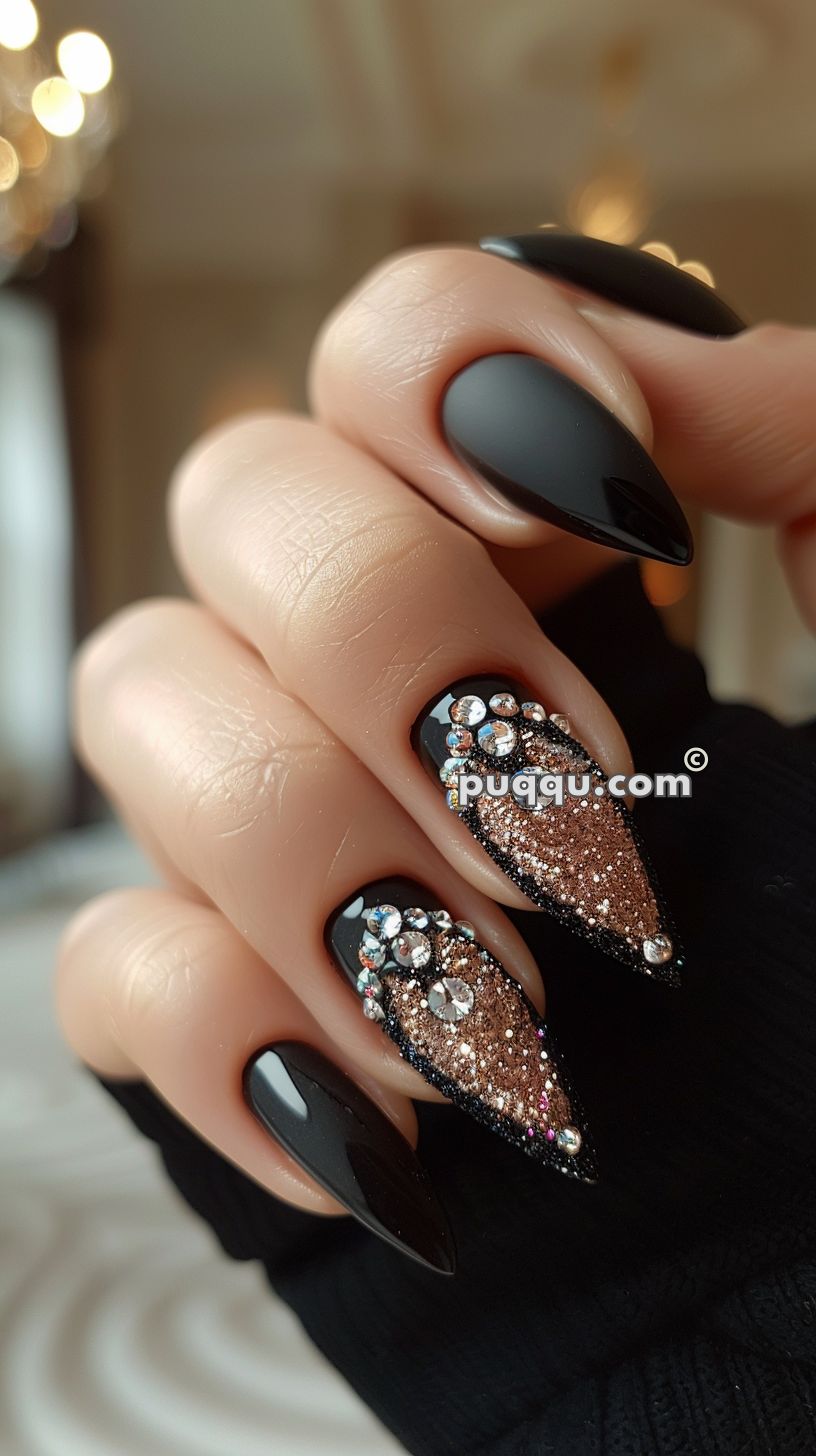 Close-up of hand with black almond-shaped nails, two featuring gold glitter and rhinestone designs.
