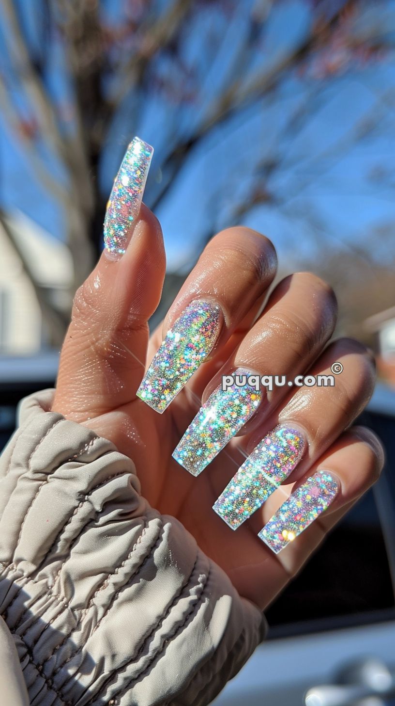 Hand with long, coffin-shaped, glittery multicolored holographic nails, held against a blurred outdoor background.