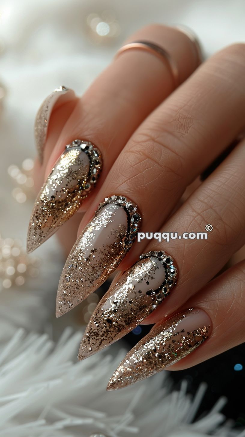 Close-up of hand with long, pointed nails painted in a sparkly gold gradient with rhinestone accents.