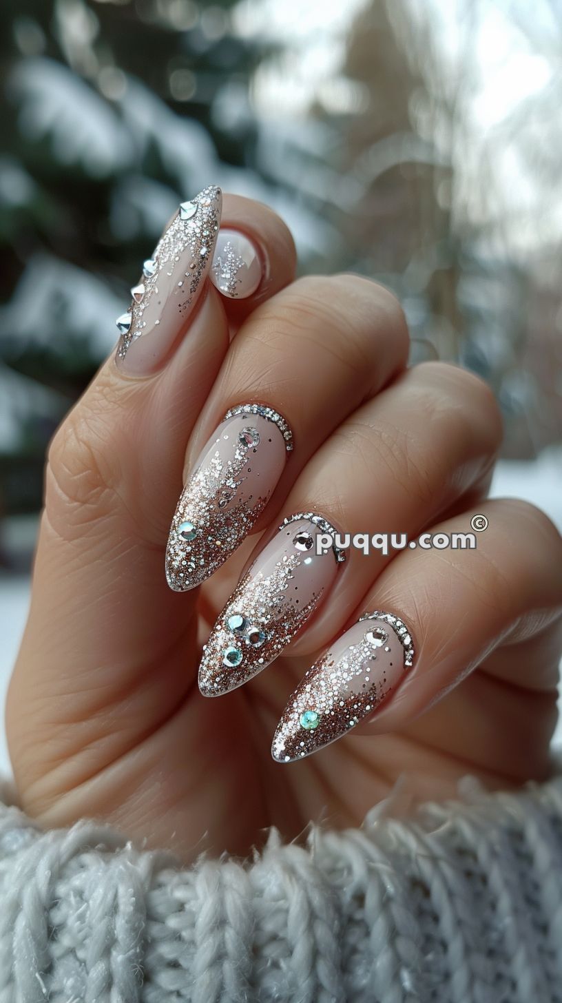 Hand with long, almond-shaped nails decorated with nude polish, silver glitter, rhinestones, and jewel accents.