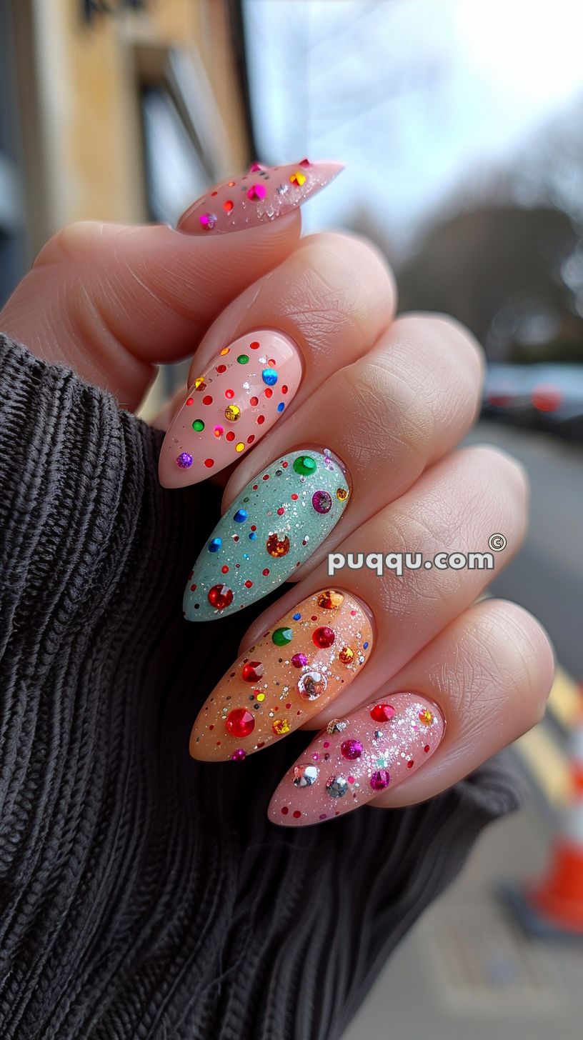 A hand with four fingernails painted in pastel colors (pink, green, and peach) with various colorful rhinestones and glitter decorations, held up against an outdoor urban background.