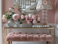Elegant entryway with a pink tufted bench, floral rug, glass table adorned with vases of flowers, floral table lamp, and a round mirror reflecting a staircase.