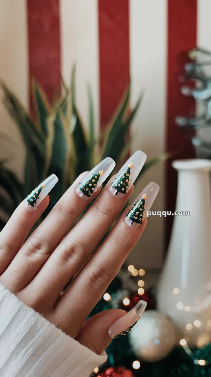 A hand with long, clear nails featuring Christmas tree nail art with gold decorations and stars.