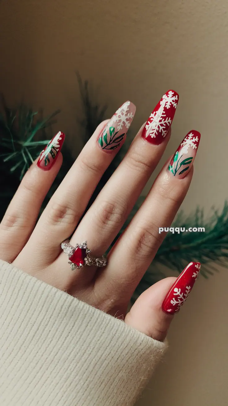 Fingers with red and white festive nail art featuring snowflakes and greenery; wearing a ring with a red gemstone.