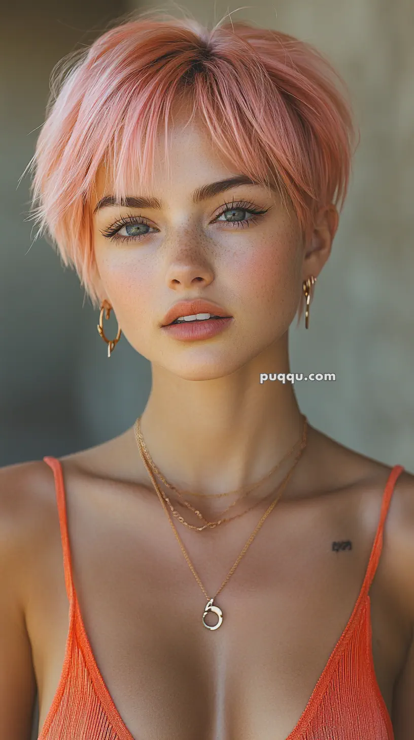 Person with short pink hair and wearing an orange top, gold hoop earrings, and layered necklaces.