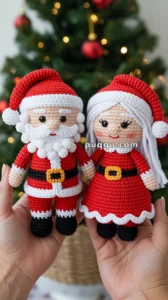 Crocheted dolls of Santa Claus and Mrs. Claus wearing red outfits with white trims, held in hands, with a Christmas tree adorned with red baubles and lights in the background.