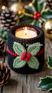 A candle in a glass holder wrapped in a knitted black cover with red accents, decorated with crocheted green leaves and red berries, surrounded by pine cones and festive holiday decorations on a wooden surface.