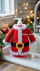 A soap dispenser dressed in a crocheted cover resembling a Santa Claus outfit, featuring red fabric with white trim and a yellow belt buckle, set on a kitchen counter with festive decorations and lights in the background.