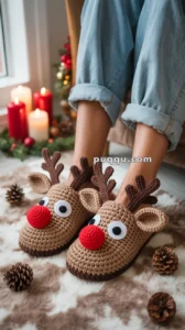 A pair of feet wearing crocheted reindeer slippers with bright red noses and antlers, surrounded by pinecones on a soft surface, with lit candles and festive decorations in the background.