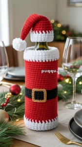 A wine bottle covered with a crocheted Santa Claus outfit, featuring a red suit with a black belt and a Santa hat, placed on a festive table setting with holiday decorations.
